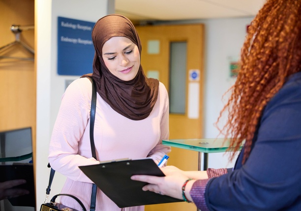 The focus is on the patient, receiving a clipboard with information on, handed by a receptionist. Both smiling and happy, ready for the patient to start their treatment at The London Clinic.