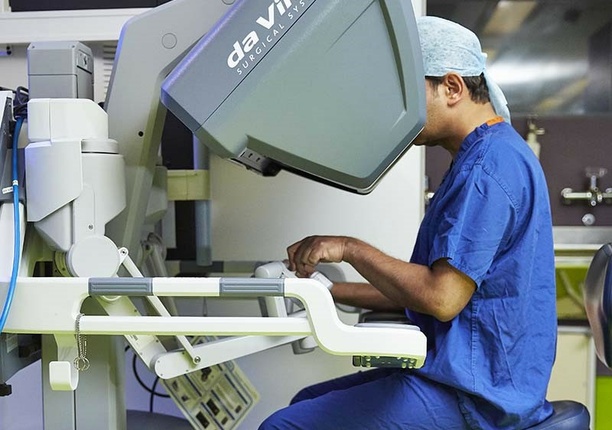 A consultant in scrubs using the da Vinci robot for surgery purposes. The consultants face is hidden as they are facing the console.
