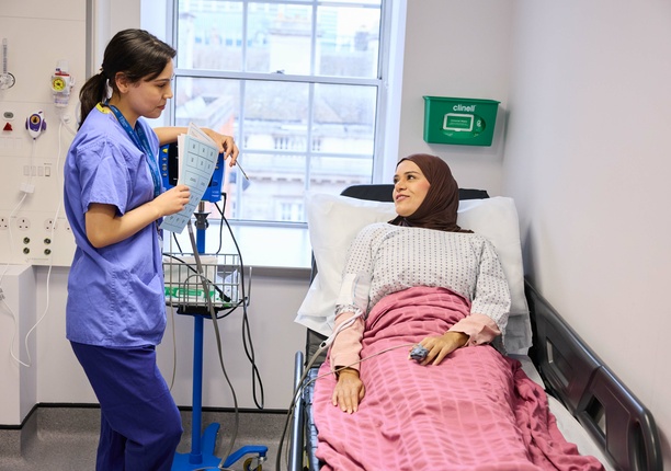 A photo of a nurse talking to a patient lying in bed, smiling at each other, whilst the nurse is taking blood pressure.