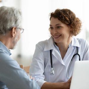 Smiling woman doctor listening to mature patient complaints at meeting in hospital, friendly therapist physician wearing white uniform with stethoscope consulting senior man at medical appointment