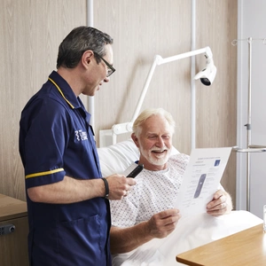 An action shot of CNS, Patrick, with an older male patient in a London Clinic hospital bed, reading a leaflet