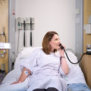 A photo of a patient lying on a hospital bed at the London Clinic using the in-house phone. The patient is connected to an IV drip and is appearing to talk on the phone.
